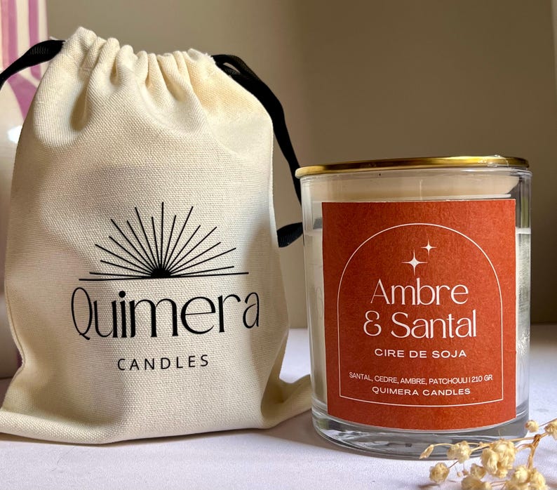 May include: A clear glass candle jar with a gold rim and a red label that reads "Ambre & Santal". The candle is next to a cream-colored drawstring bag with the "Quimera Candles" logo. The candle is made with soy wax.