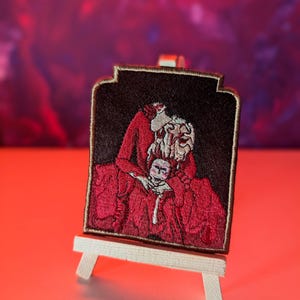 Peut inclure: Patch brodé représentant deux personnages enlacés en rouge et blanc. Le patch est encadré d'une bordure brune et est présenté sur un petit chevalet en bois.
