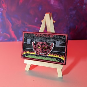 Könnte beinhalten: Gestickter Aufnäher mit einem roten Teufel mit gelben Augen und einem finsteren Grinsen. Der Aufnäher hat einen schwarzen Rand und ein grünes und gelbes Flammendesign. Er wird auf einer kleinen Holzstaffelei vor farbenfrohem Hintergrund präsentiert.