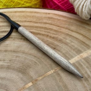 May include: A close-up of a wooden knitting needle with a black tip, lying on a wooden surface. The needle is light brown in color and has a smooth finish.