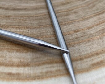 May include: Two silver knitting needles, one lying on top of the other, on a wooden surface.