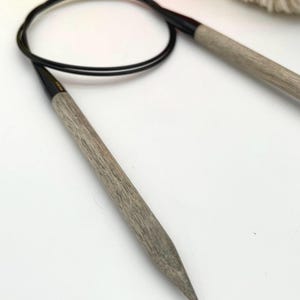 May include: A pair of gray knitting needles with black tips. The needles are lying on a white surface.