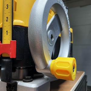 May include: A yellow and silver power tool adjustment mechanism featuring a circular silver handwheel with a yellow knob. A vertical yellow ruler with black markings is visible on the left, showing measurements in inches.