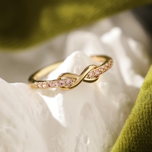 May include: A gold infinity ring with pink gemstones. The ring is delicate and features a simple design.