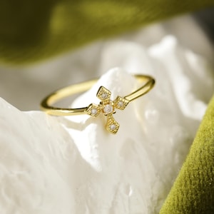 Pode incluir: Um anel de ouro com um pequeno design de cruz adornado com pedras de zircônia cúbica transparentes.