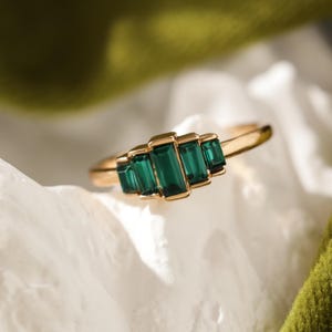 Op de afbeelding: Een gouden ring met vijf smaragdgroene edelstenen met smaragdgroene slijpsel. De edelstenen zijn in een horizontale rij geplaatst, met een gouden band. De ring wordt gepresenteerd op een wit, getextureerd oppervlak.