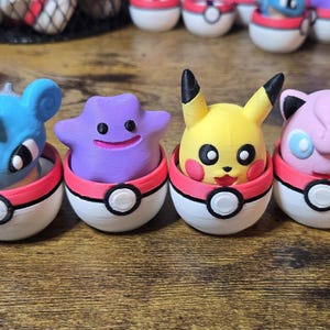 May include: Four colorful Pokémon figures, including Pikachu, Ditto, and Jigglypuff, sit inside red and white Poké Ball containers. The figures are blue, purple, yellow, and pink, with black and white accents. The items are on a wooden surface.