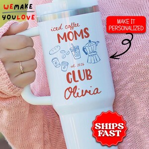 May include: White insulated tumbler with a handle, featuring the text "iced coffee MOMS CLUB Olivia" in red and blue. The design includes coffee-related graphics. The tumbler has a silver lid and is held by a person.
