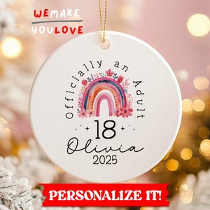 May include: White ceramic ornament with a rainbow design and the text "officially an Adult 18 Olivia 2025". The ornament has a gold string for hanging and is decorated with floral accents. The words "We Make You Love" and "Personalize It!" are also visible.