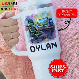 May include: White insulated tumbler featuring a gaming setup design with a green and black gaming chair, computer, and the name "DYLAN" in pixelated letters. The tumbler also has the text "WE MAKE YOU LOVE".