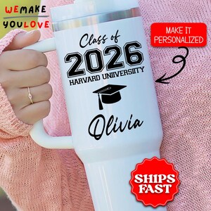 May include: White insulated tumbler with a handle, featuring the text "Class of 2026 Harvard University" and the name "Olivia". A graduation cap graphic is below the university name. The tumbler has a silver top and a red sticker that says "Make it Personalized".