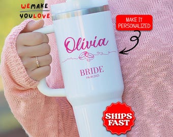 May include: White insulated tumbler with a handle, featuring the name "Olivia" in pink script, along with wedding rings and the word "BRIDE" and a date. The tumbler has a silver rim and a white handle. The text "WE MAKE YOU LOVE" is visible.