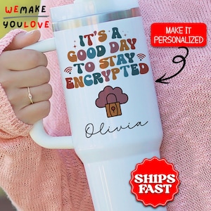May include: A white insulated tumbler with a handle, featuring the text "IT'S A GOOD DAY TO STAY ENCRYPTED" in colorful letters. A cloud with a lock graphic is below the text, with the name "Olivia" below that. A red sticker says "MAKE IT PERSONALIZED".