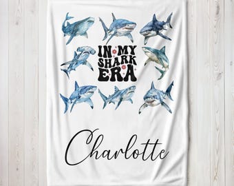May include: White blanket featuring watercolor illustrations of various sharks. The phrase "IN MY SHARK ERA" is centered, with the name "Charlotte" below. The blanket has a soft, cozy appearance.