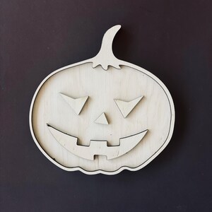 May include: A white wooden pumpkin with a carved jack-o'-lantern face. The pumpkin has a wide smile, two triangular eyes, and a small triangular nose.