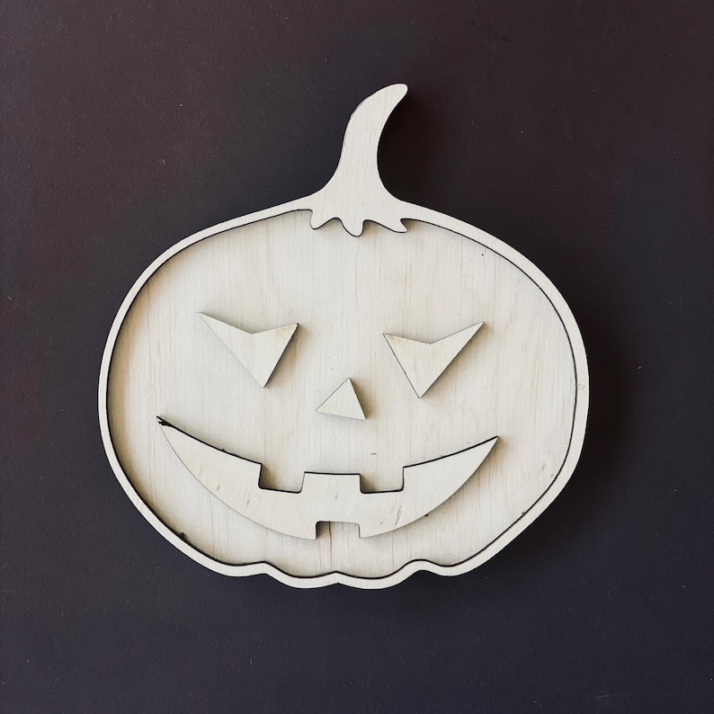 May include: A white wooden pumpkin with a carved jack-o'-lantern face. The pumpkin has a wide smile, two triangular eyes, and a small triangular nose.