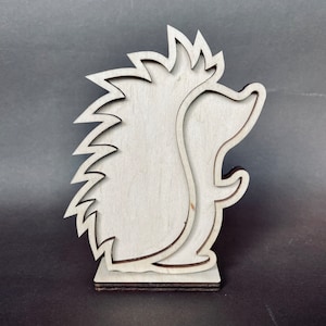 May include: A wooden cutout of a hedgehog with a simple design. The hedgehog is standing on a small base.