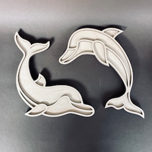 May include: Two wooden dolphin wall decor pieces, one facing left and the other facing right. The dolphins are cut out of wood and have a simple, elegant design.