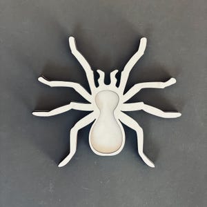 May include: A white wooden spider cutout with a black outline. The spider has eight legs and a round body.