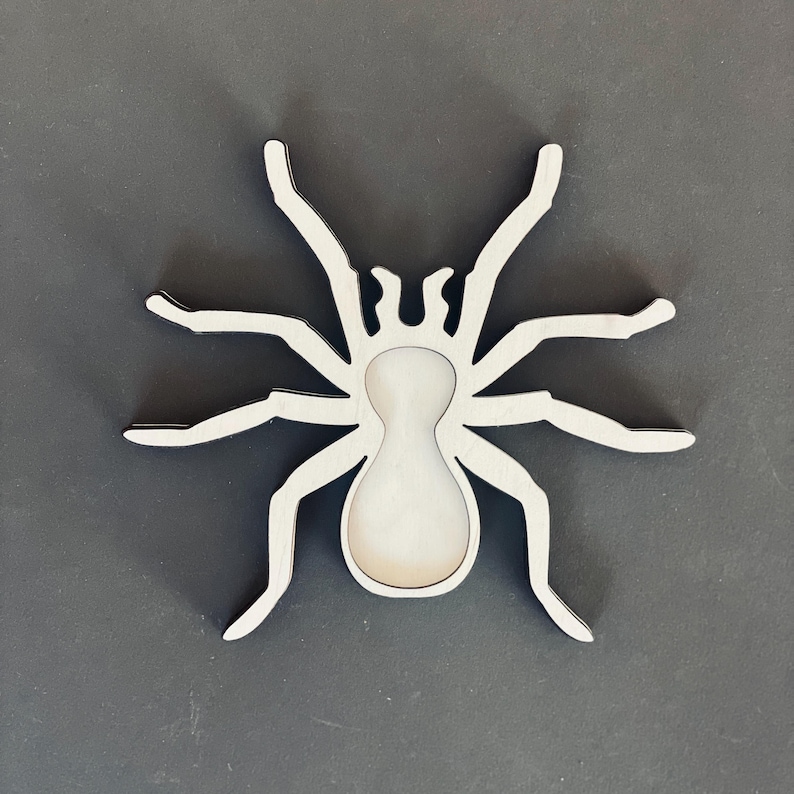 May include: A white wooden spider cutout with a black outline. The spider has eight legs and a round body.