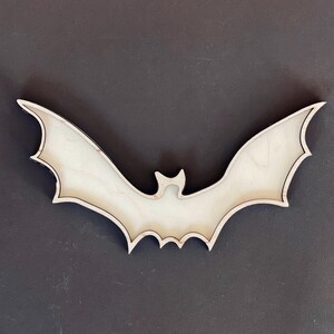 May include: A wooden bat cutout, painted white, with a black outline. The bat is shaped like a traditional Halloween bat with wings spread wide.