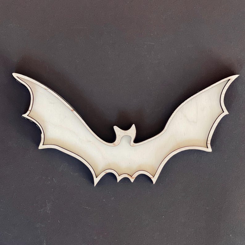 May include: A wooden bat cutout, painted white, with a black outline. The bat is shaped like a traditional Halloween bat with wings spread wide.