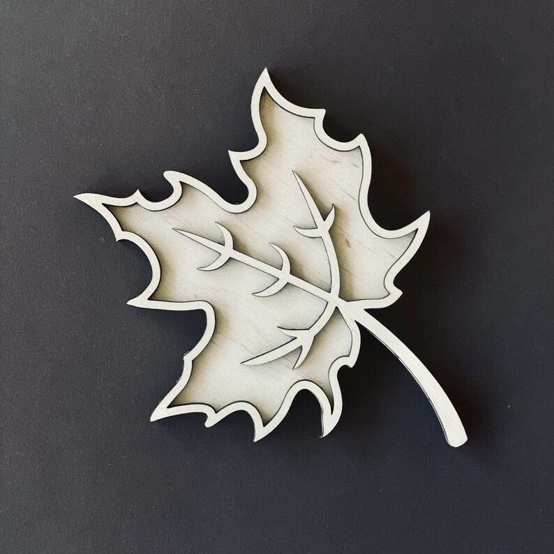 May include: A white wooden maple leaf cutout with a detailed vein pattern. The leaf is a simple, elegant design that would look great in a home decor setting.