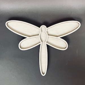 May include: A wooden dragonfly wall decor with a cutout design. The dragonfly is light brown and has a detailed body and wings.