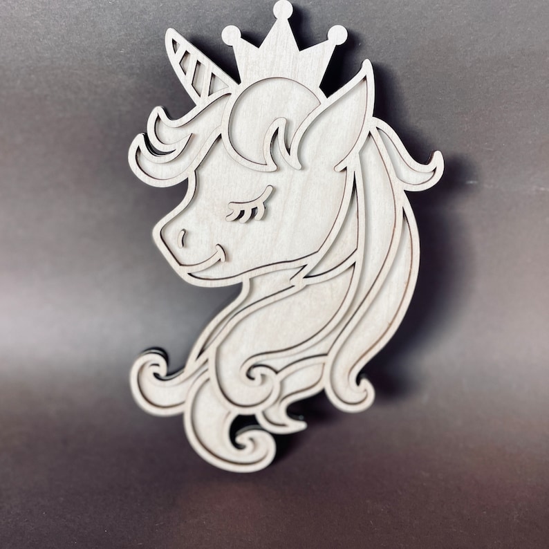 May include: A wooden cutout of a unicorn head with a crown. The unicorn has closed eyes and a detailed mane.