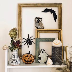 May include: A shelf with a variety of Halloween decorations, including a mosaic owl, a bat, a spider, a pumpkin, a ghost, and a jack-o'-lantern. The decorations are displayed in front of a gold picture frame.