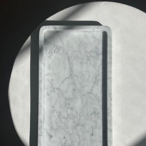 May include: A white marble rectangular tray with a raised edge. The tray is sitting on a white surface.