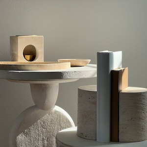 K&ouml;nnte beinhalten: Ein minimalistisches Stillleben-Arrangement. Ein kleiner, runder Tisch tr&auml;gt eine Sammlung von Objekten: einen quadratischen Block mit einer kreisf&ouml;rmigen &Ouml;ffnung, eine kleine Messingschale und mehrere steinartige Zylinder und rechteckige Formen. Das Farbschema ist &uuml;berwiegend neutral.