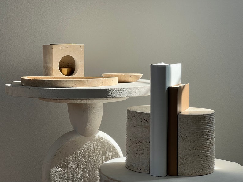 K&ouml;nnte beinhalten: Ein minimalistisches Stillleben-Arrangement. Ein kleiner, runder Tisch tr&auml;gt eine Sammlung von Objekten: einen quadratischen Block mit einer kreisf&ouml;rmigen &Ouml;ffnung, eine kleine Messingschale und mehrere steinartige Zylinder und rechteckige Formen. Das Farbschema ist &uuml;berwiegend neutral.