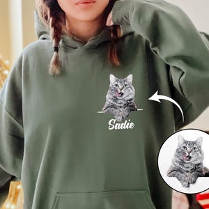 May include: A green hooded sweatshirt features a custom design. A gray cat image is displayed on the front pocket area with the name Sadie printed below.  A circular inset shows an additional image of the same cat. The sweatshirt is a comfortable, oversized style, perfect for pet lovers.