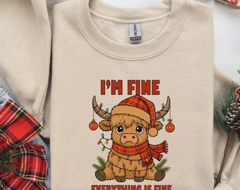 May include: Beige sweatshirt with a graphic of a Highland cow wearing a plaid hat and scarf, decorated with Christmas lights and ornaments. The text "I'M FINE EVERYTHING IS FINE" is printed above and below the cow.