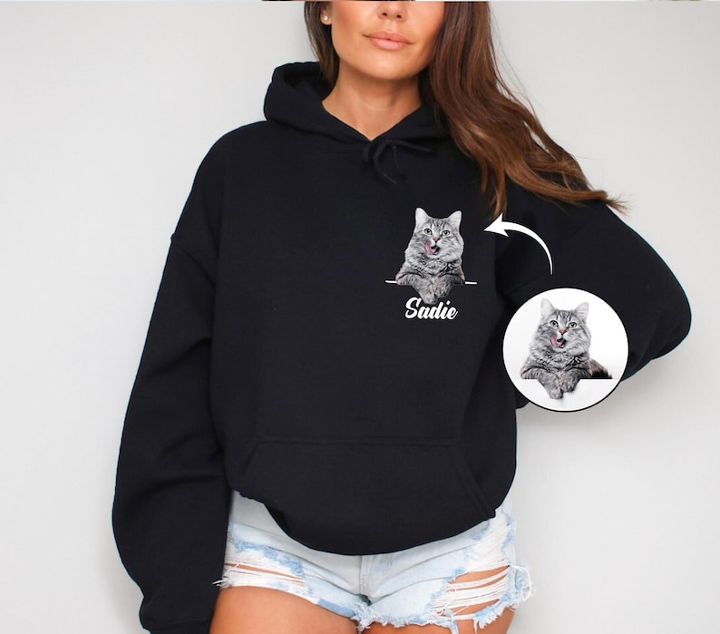 Puede incluir: Sudadera con capucha negra con un retrato de gato personalizado. El gato es gris y blanco, con la lengua fuera. El nombre "Sadie" est&aacute; impreso debajo del gato. Tambi&eacute;n hay una imagen circular m&aacute;s peque&ntilde;a del gato.