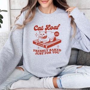 May include: Gray sweatshirt with a red graphic of a cat sitting on a cutting board. The graphic says "Cat Loaf" and "Freshly Baked Just For You".
