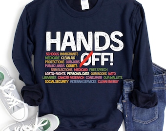 May include: Navy blue sweatshirt with the text "HANDS OFF!" in white. The text is surrounded by a list of issues in colorful text, including schools, immigrants, medicare, clean air, protections, our jobs, public lands, courts, fair elections, medicaid, free speech, LGBTQ+ rights, personal data, our books, NATO, libraries, cancer research, consumer, our wallets, social security, veteran services, and clean energy.