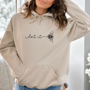 May include: Beige hoodie with a front pocket and drawstring. The hoodie features the phrase "let it" in a cursive font, connected to a stylized bee graphic. The bee is black and white, with detailed wings. The overall design is simple and elegant.
