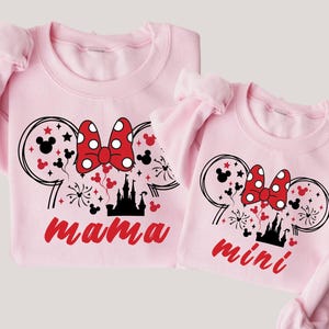 May include: Pink sweatshirts with a black and white illustration of Mickey Mouse ears with a red bow and a black castle. The larger sweatshirt says "mama" and the smaller sweatshirt says "mini".