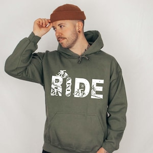 May include: An olive green hoodie with the word "RIDE" in large white block letters. The letters are filled with white illustrations of skiers and snowboarders. The person in the image is wearing a brown beanie.
