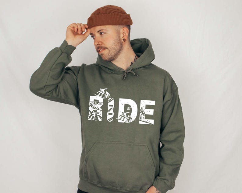 May include: An olive green hoodie with the word "RIDE" in large white block letters. The letters are filled with white illustrations of skiers and snowboarders. The person in the image is wearing a brown beanie.