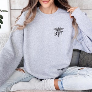May include: Light gray sweatshirt with a black caduceus symbol and the letters "R T" printed on the left chest. The sweatshirt has long sleeves and a crew neck. The fabric appears to be a soft, comfortable material.