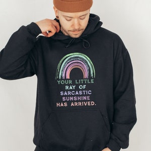 Puede incluir: Sudadera con capucha negra con un gr&aacute;fico de arco&iacute;ris y el texto "Your little ray of sarcastic sunshine has arrived."