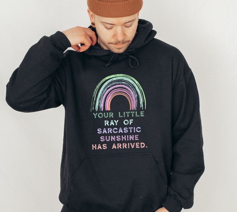 Puede incluir: Sudadera con capucha negra con un gr&aacute;fico de arco&iacute;ris y el texto "Your little ray of sarcastic sunshine has arrived."