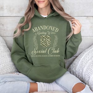May include: Olive green hoodie with the text "Abandoned Hunting Wives Social Club" in a decorative font. The hoodie has a front pocket and a drawstring hood. The text includes the phrase "Hello Hunting Season, Goodbye Husband."