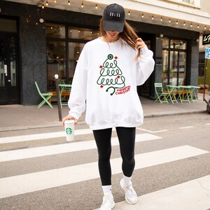 Pu&ograve; includere: Una felpa bianca con un disegno di albero di Natale verde e le parole "MERRY CHRISTMAS" in rosso. La persona indossa un berretto da baseball nero con "11:11", leggings neri e scarpe da ginnastica bianche.