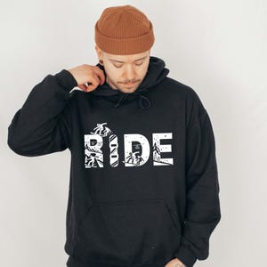 May include: Black hoodie with the word "RIDE" in white block letters. The letters are filled with images of skiers and snowboarders. The hoodie has a front pocket and a drawstring hood.