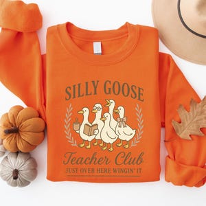 May include: An orange sweatshirt featuring the text "Silly Goose Teacher Club" and a graphic of five cartoon geese. One goose wears glasses and holds a book, another holds a coffee cup, and one wears a bow. The phrase "Just Over Here Wingin' It" is below the graphic.