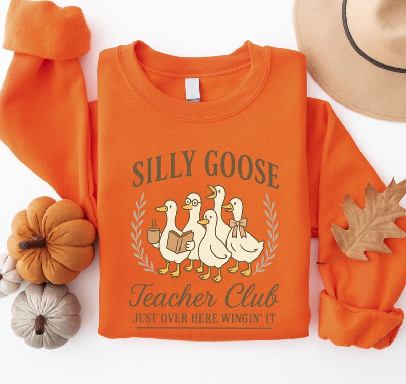 May include: An orange sweatshirt featuring the text "Silly Goose Teacher Club" and a graphic of five cartoon geese. One goose wears glasses and holds a book, another holds a coffee cup, and one wears a bow. The phrase "Just Over Here Wingin' It" is below the graphic.
